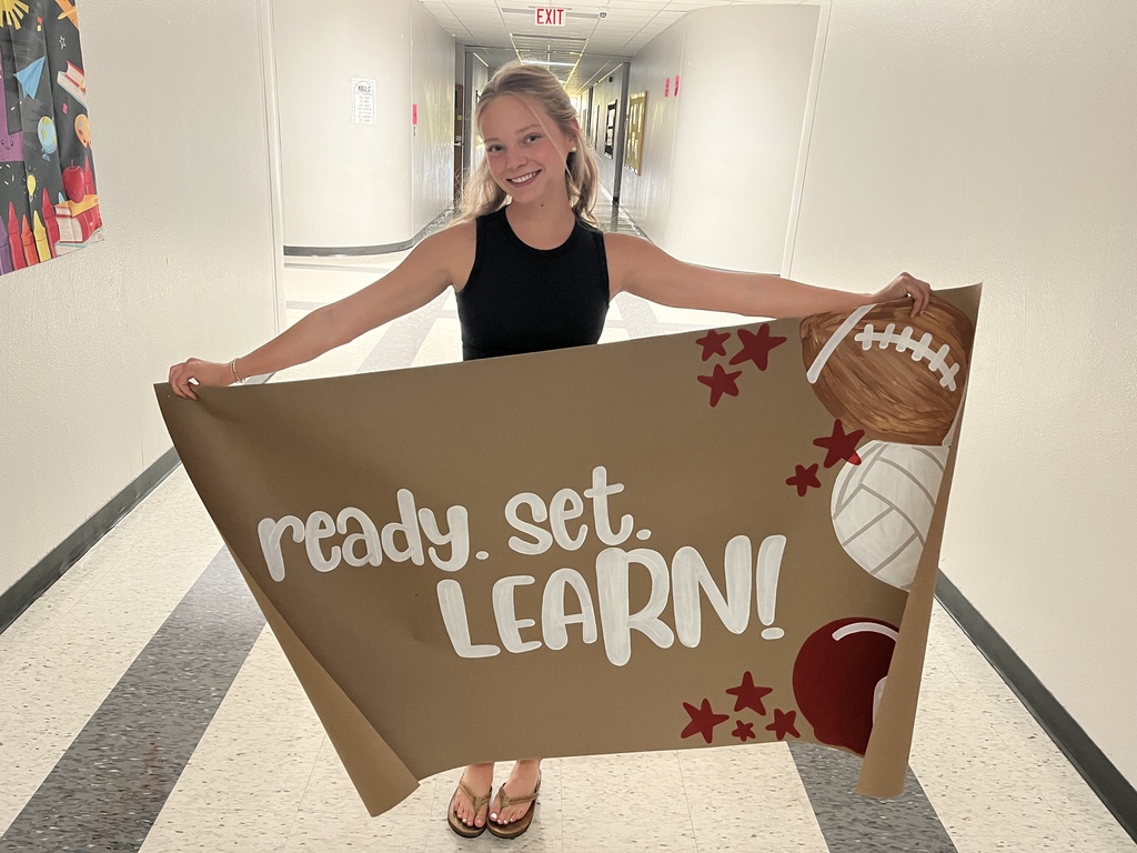 Smiling Russell standing in a school hallway holding up a large brown banner that reads "ready. set. LEARN!" decorated with painted footballs, a volleyball, stars, and maroon accent details.