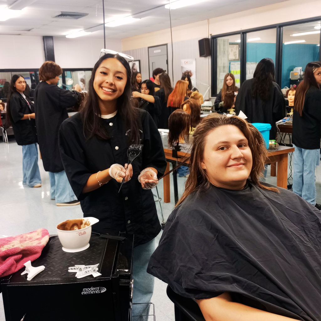 Cosmetology student Abby smiling while applying hair color to client Payton Baklik in a classroom salon setup.