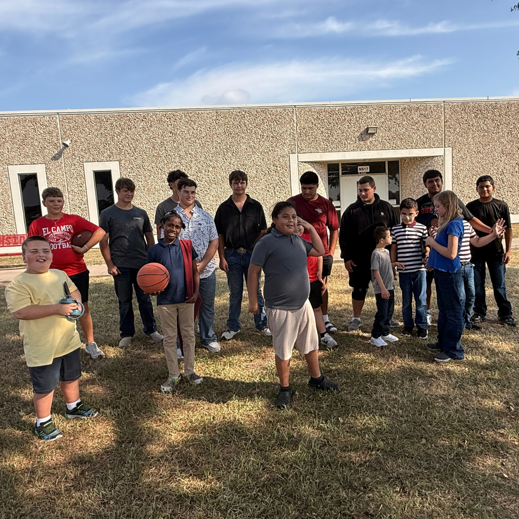 A group of elementary students and high school boys smiling together outside near a school building, with one student holding a basketball.