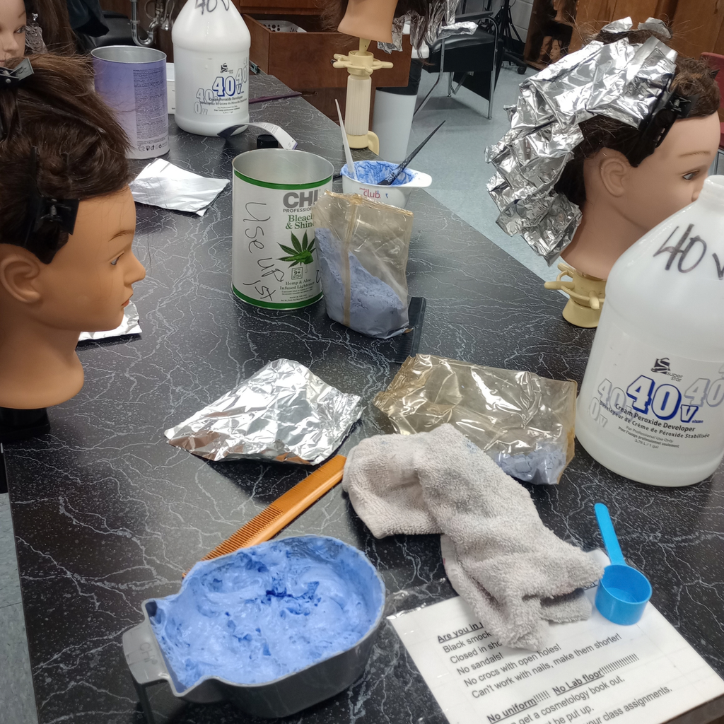 Cosmetology setup featuring mannequin heads, hair foil wraps, blue bleach mix in a bowl, and hair developer bottles on a table.