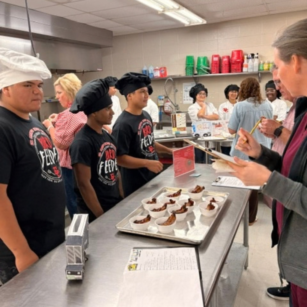 Culinary arts students wearing black "Bird Feeder" t-shirts and chef hats stand behind a stainless steel kitchen counter, serving ribs on a tray to a judge holding a clipboard.