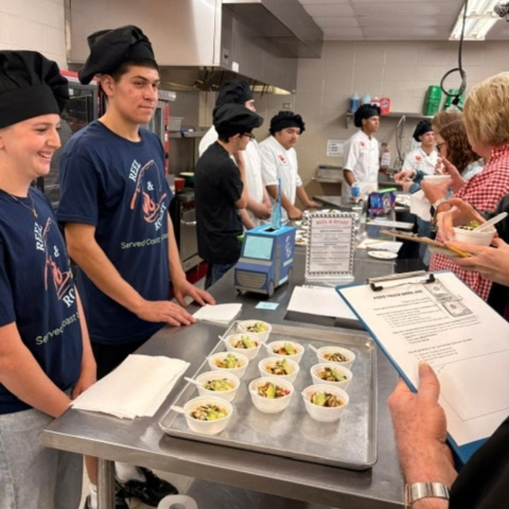 Culinary arts students serve samples of fish taco bowls from behind a kitchen counter to judges holding clipboards, while small tasting bowls are arranged on a tray in the foreground.