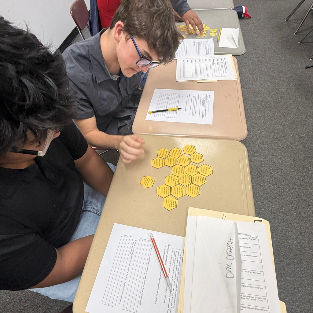 At works with a classmate at a desk to arrange yellow hexagonal cards during a classroom activity.