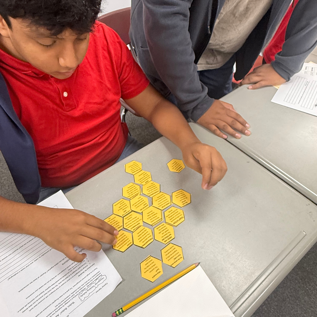 A student in a red shirt works with classmates at a desk to arrange yellow hexagonal cards during a classroom activity.