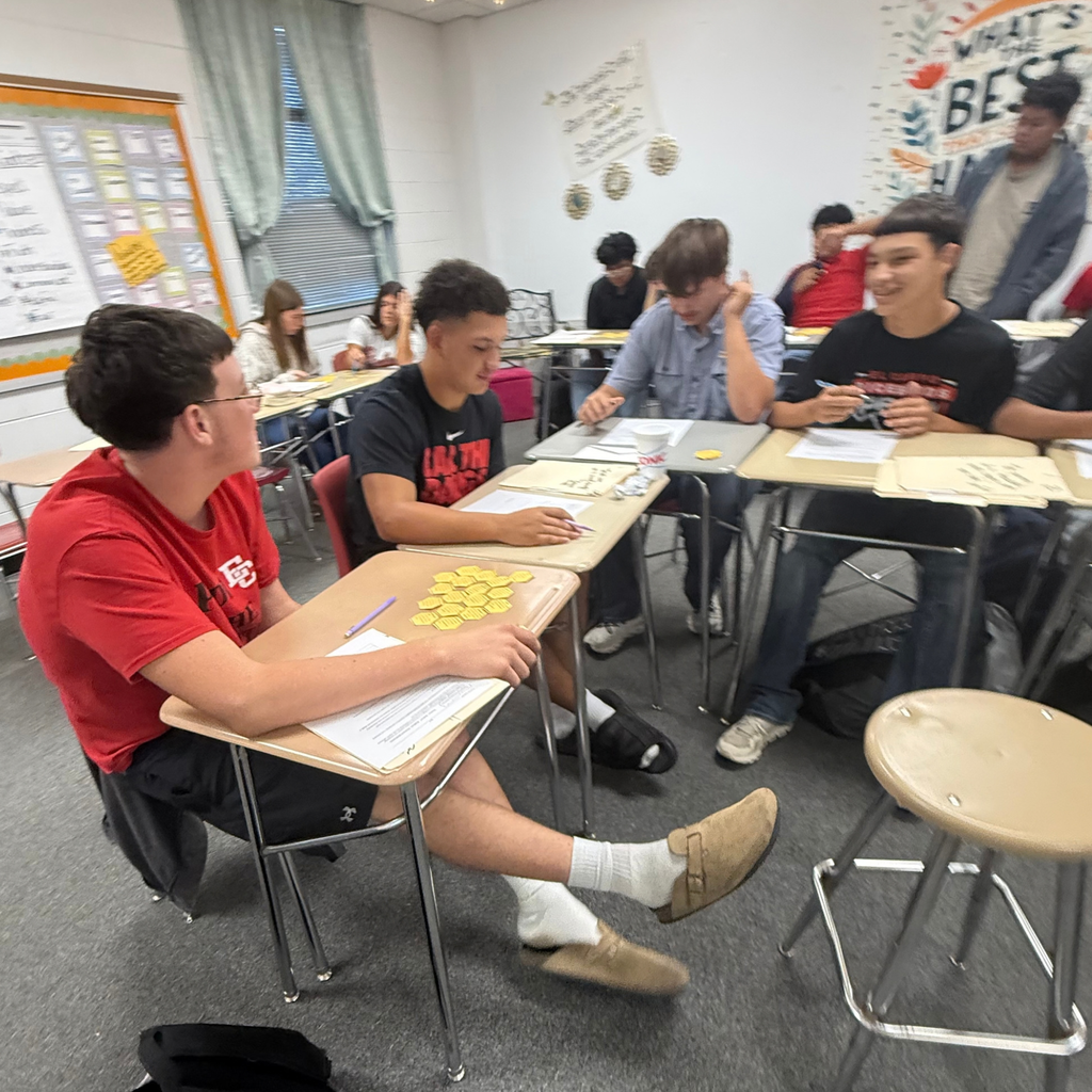 A student in a red shirt works with classmates at a desk to arrange yellow hexagonal cards during a classroom activity.