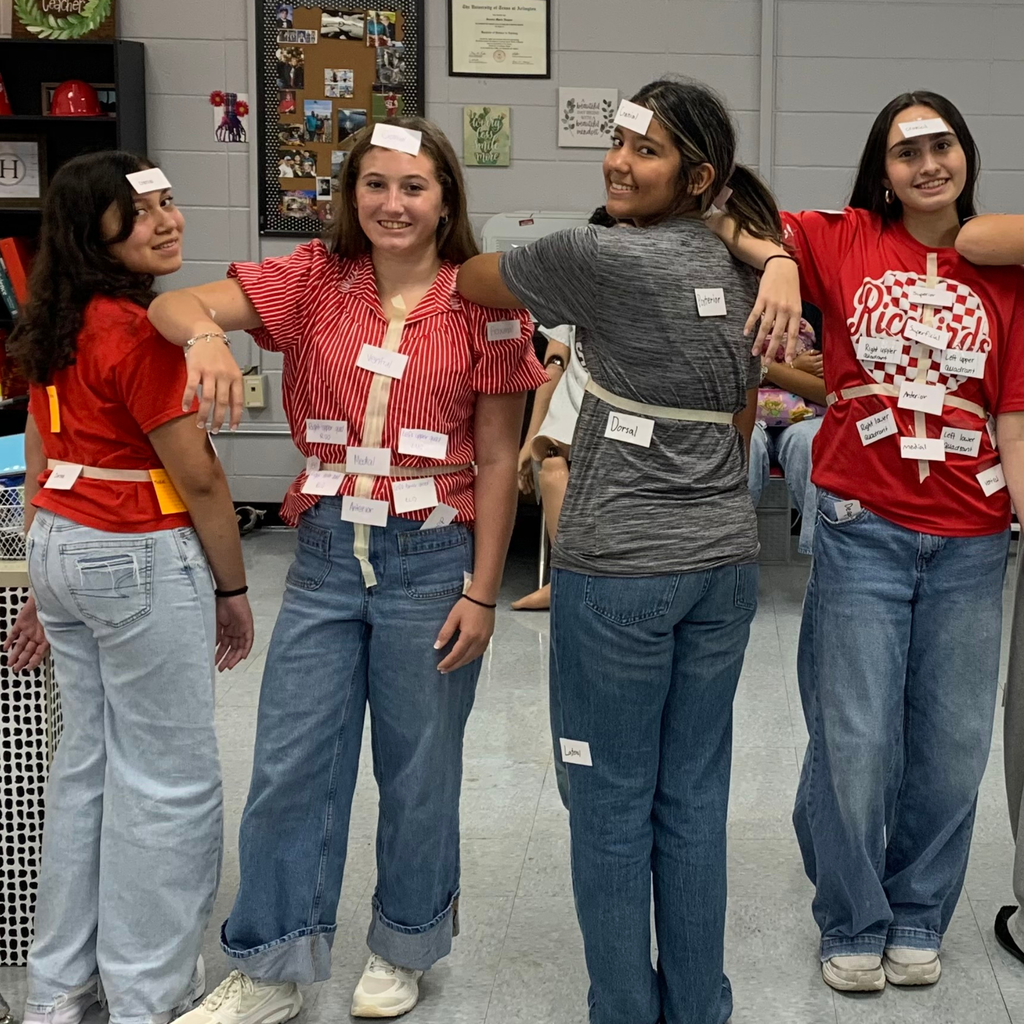 Four high school students in a classroom pose together while wearing labels on their clothing to identify directional body terms during a health science activity.