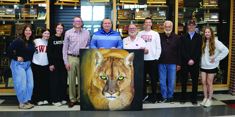 students and administartive staff stand around the painting with artist Milo West in front of a trophy case