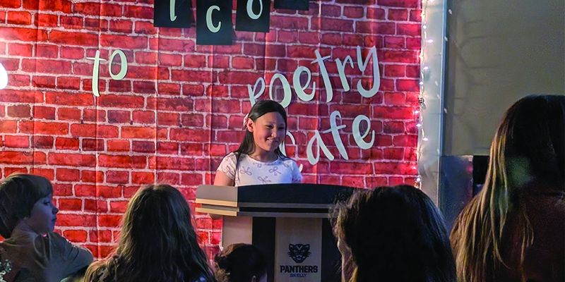 A students smiles behind a podium in front of a backdrop that says Welcome to Poetry Café