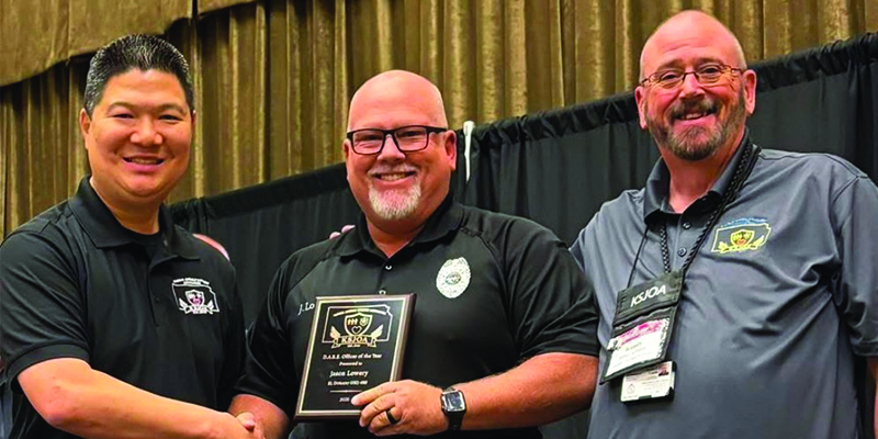 Jason Lowery shakes hands with a Kansas Juvenile Officers Association official while holding a plaque announcing him as 2026 DARE Officer of the Year while another man stands to Lower's other side