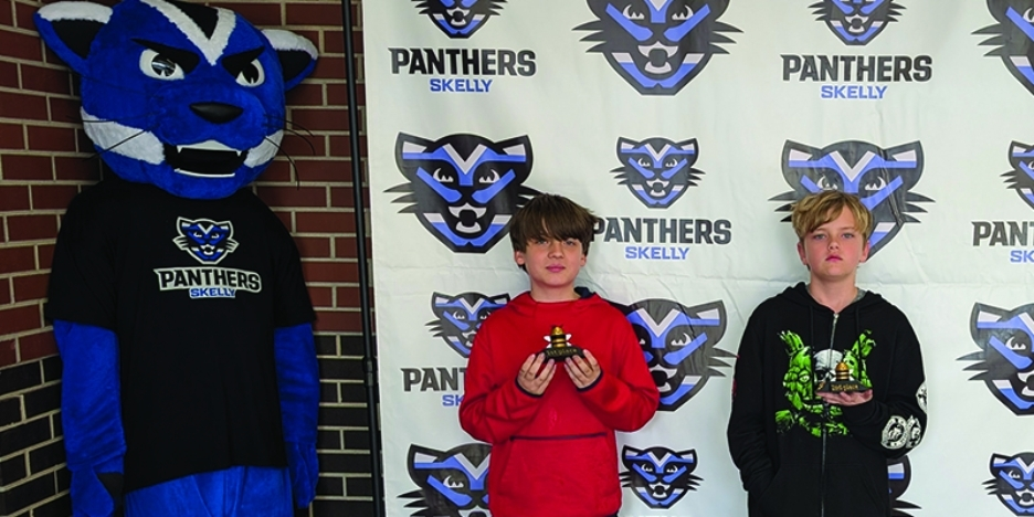 Skelly spelling bee winner and runner-up pose in front of the Skelly backdrop with the panther mascot, holding their trophies