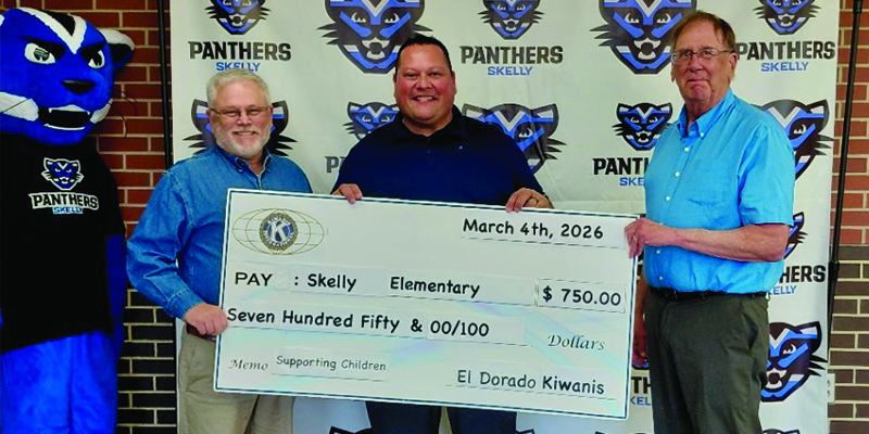 Three men hold a check from Kiwanis to Skelly Elementary for $750, standing in front of a Skelly logo backdrop
