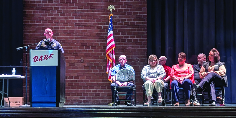 Police Chief Mike Holton stands at a podium while Sheriff Monty Hughey and USD 490 SROs and admin are seated on the side of the stage