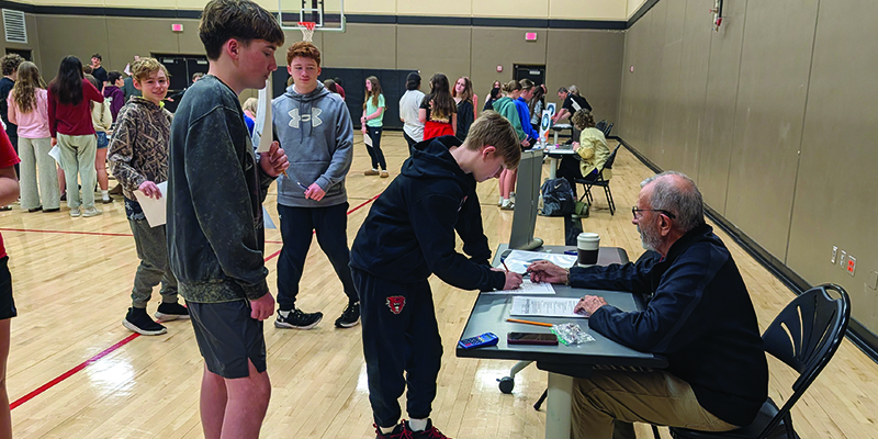 a student works on something at a table with an adult while other students wait in line