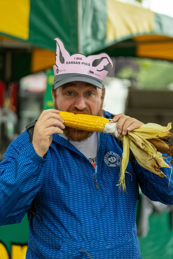 Man in a blue quarter zip eating an ear of corn with a paper hat that says "I Dig Kansas Pigs"