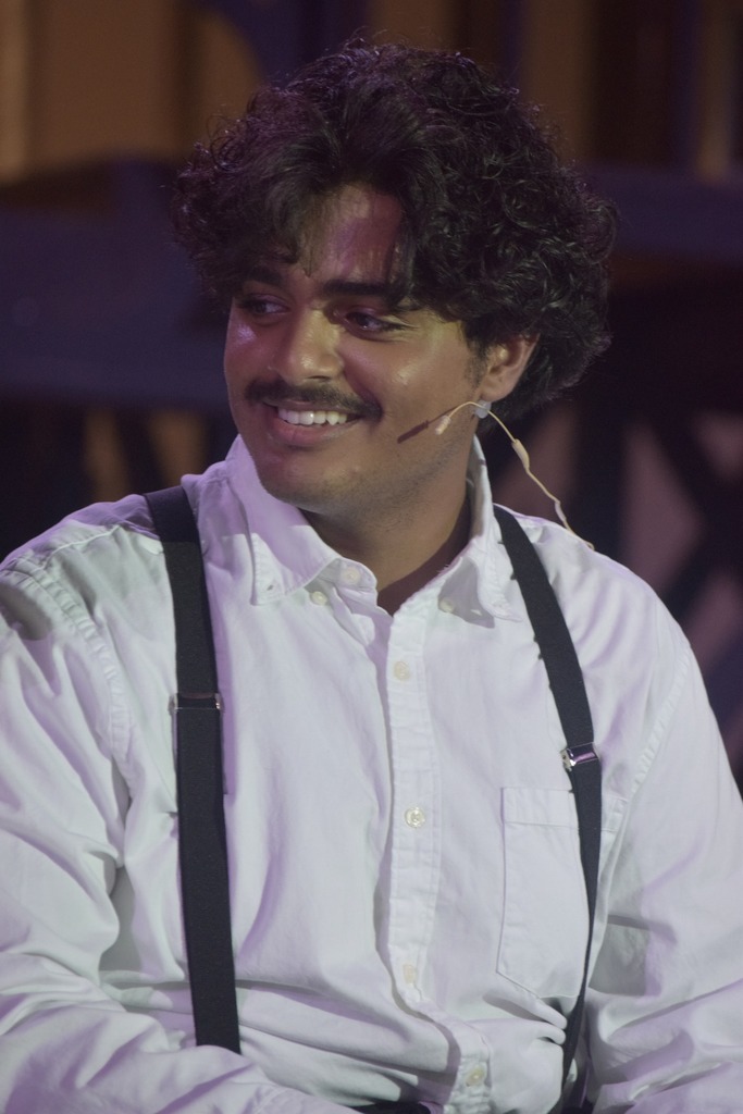 A student actor wearing a white shirt, suspenders, and a stage microphone smiles during a dress rehearsal scene from Radium Girls.