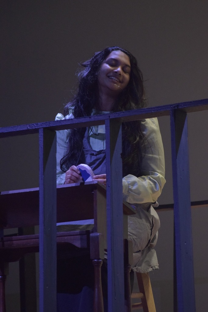 A student actor in costume smiles while seated behind a stage railing during a dress rehearsal for Radium Girls.