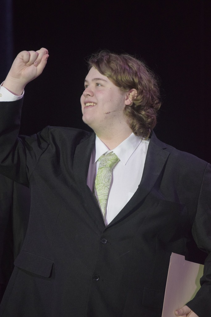 A student actor wearing a black suit and green tie smiles while raising his hand during a scene from Radium Girls.