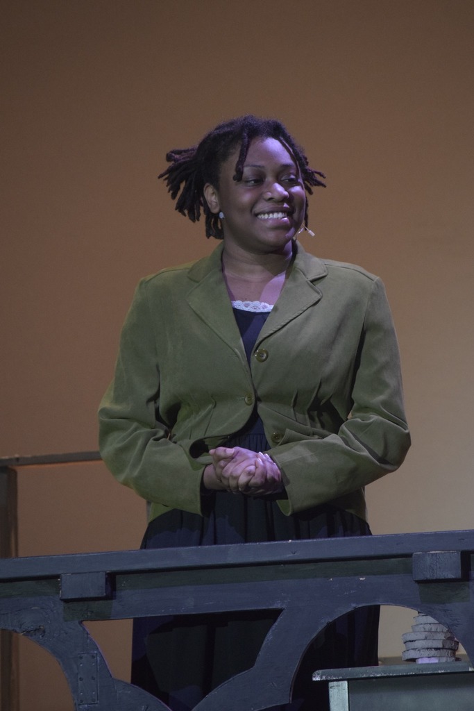 A student actor in a green jacket smiles from behind a stage railing during the EHS dress rehearsal of Radium Girls.