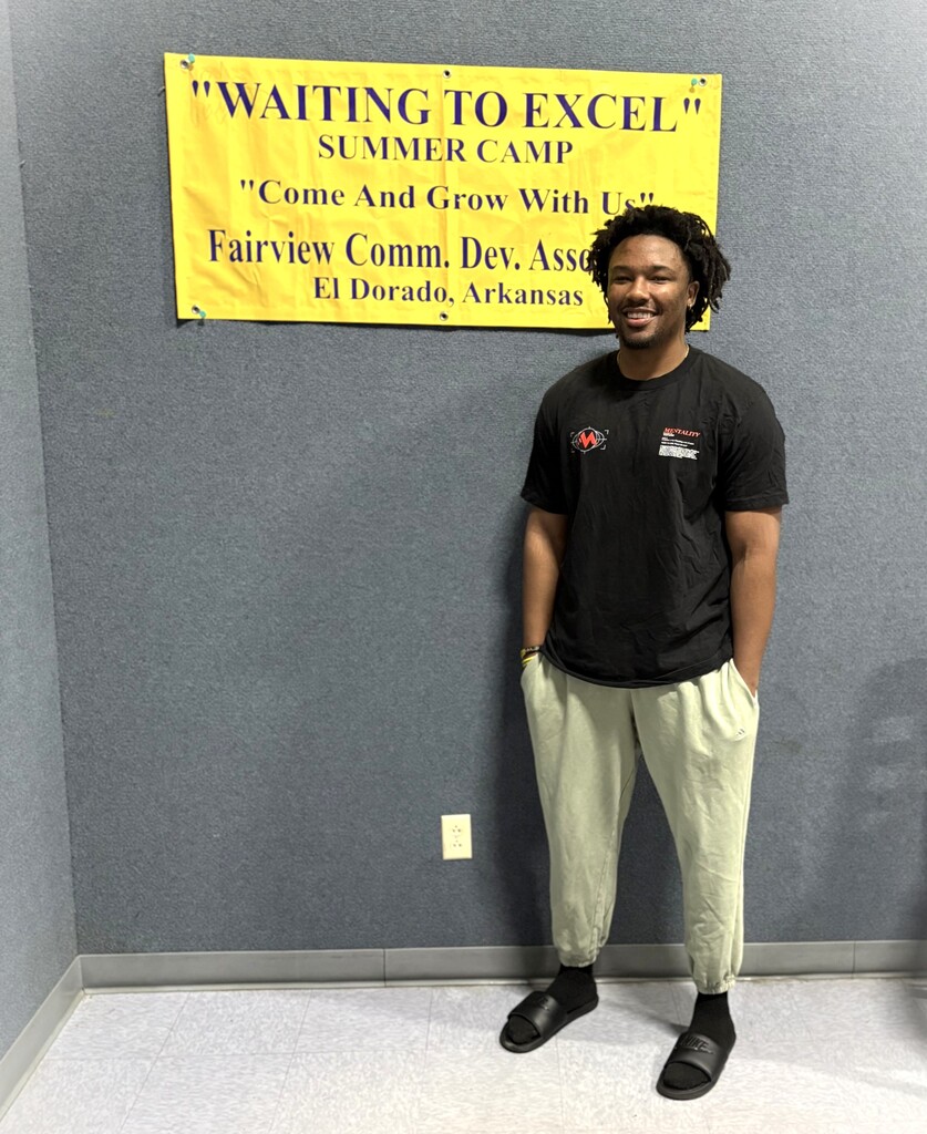 A smiling young man stands beneath a yellow “Waiting to Excel” Summer Camp banner for the Fairview Community Development Association in El Dorado, Arkansas.
