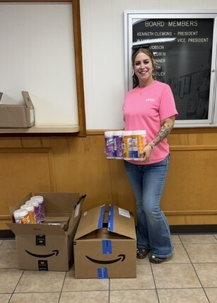 A woman in a pink T-shirt holds several canned back to school items while standing beside open donation boxes filled with food supplies.