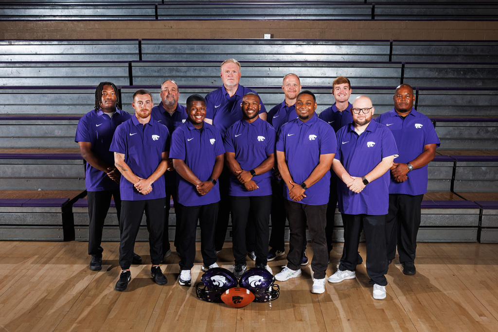 El Dorado High School Wildcats football coaching staff posing together in the gym, wearing matching purple Wildcats polo shirts. Two football helmets and a football are placed on the floor in front of the group.