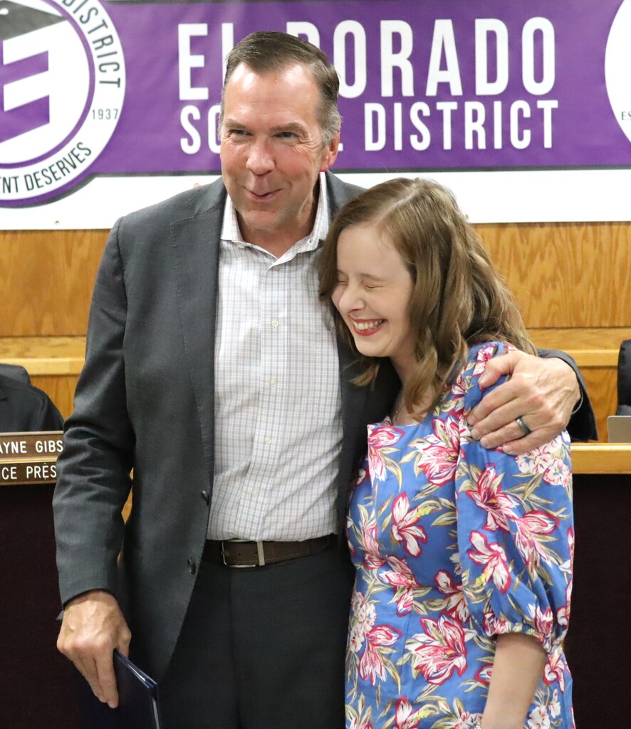 A male El Dorado School District administrator smiles with his arm around a woman in a blue floral dress during the School Board meeting. The district banner is visible behind them.