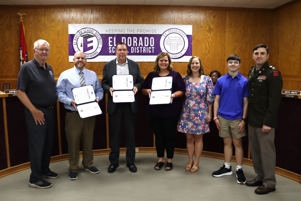 Three El Dorado School District administrators hold their ESGR Patriot Award certificates during the School Board meeting. They are joined by ESGR representatives, an EHS teacher, her family and a uniformed service member in front of the district banner.