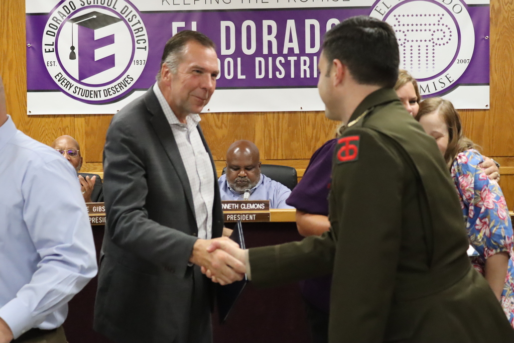 A man in a suit shakes hands with a uniformed service member during an El Dorado School District event, while district leaders and attendees stand nearby in front of district and El Dorado Promise signage.