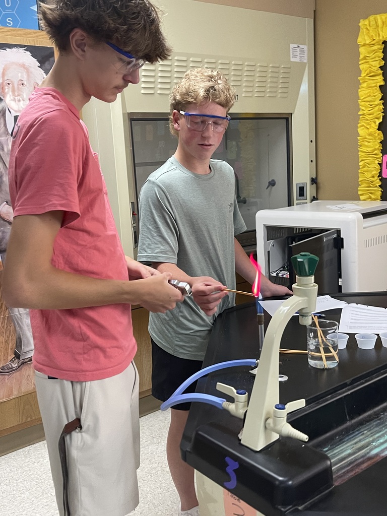 Two Advanced Chemistry students wearing safety goggles observe a bright pink-red flame produced during a flame test experiment at a lab station.
