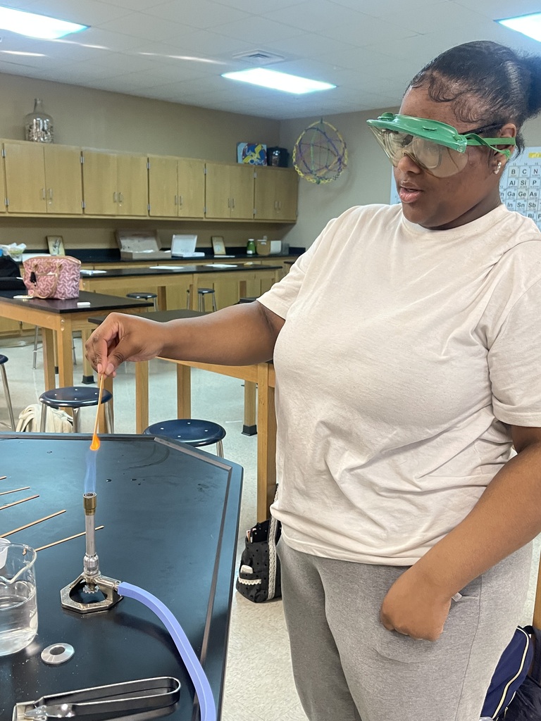An Advanced Chemistry student wearing protective goggles holds a sample over a Bunsen burner, producing a bright orange flame during a classroom flame test lab.