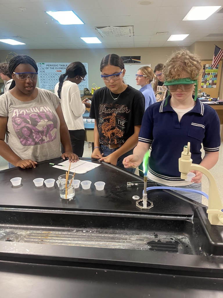 Three Advanced Chemistry students wearing protective goggles work together at a lab station as one student observes a vivid green flame during a flame test experiment.