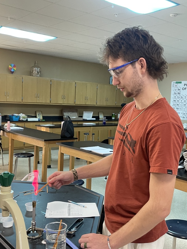 An Advanced Chemistry student wearing safety goggles observes a vivid pink-red flame during a flame test experiment in the chemistry classroom.