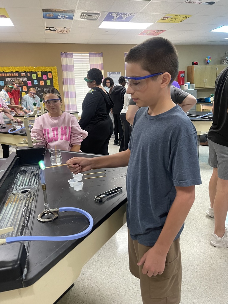 An Advanced Chemistry student wearing safety goggles holds a wooden applicator over a Bunsen burner and observes a bright green flame during a flame test lab.