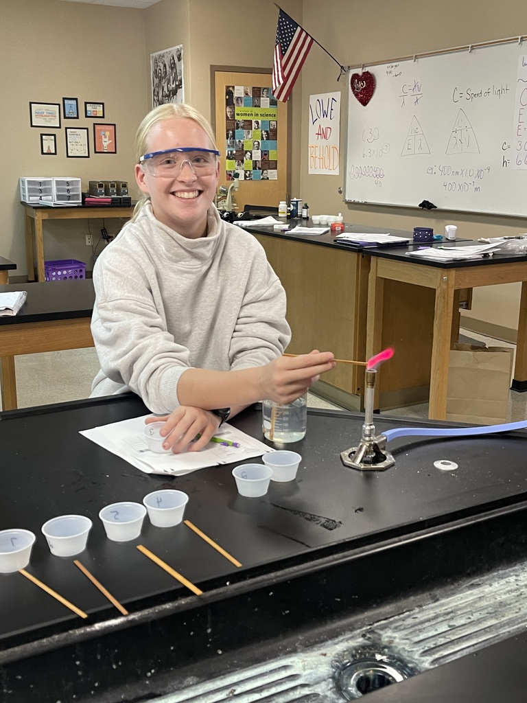 An Advanced Chemistry student smiles while holding a sample over a Bunsen burner, producing a bright pink flame during a flame test lab.
