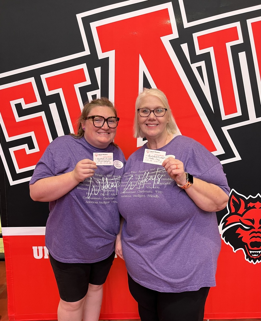 1. Heather Craig and Allison Applegate smile while holding their Special Olympics Arkansas basketball and pickleball certification cards in front of an Arkansas State University backdrop.