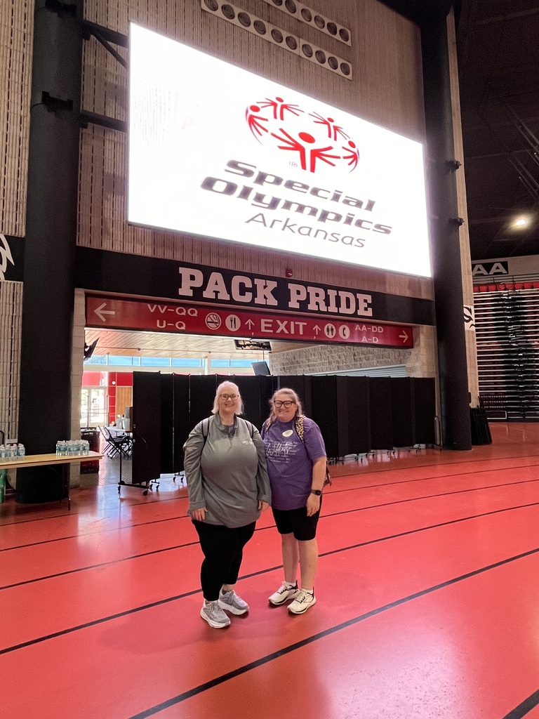 Two El Dorado School District educators pose together inside First National Bank Arena beneath a large screen displaying the Special Olympics Arkansas logo.