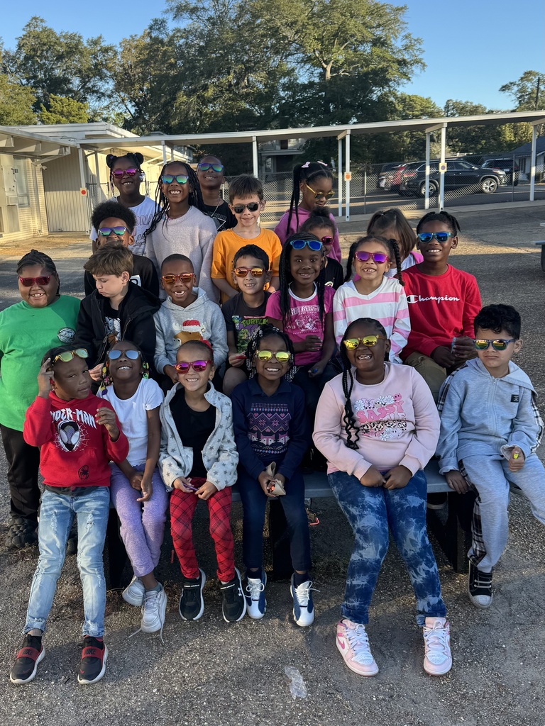 Several children in sunglasses pose for a group photo in a parking lot with trees.