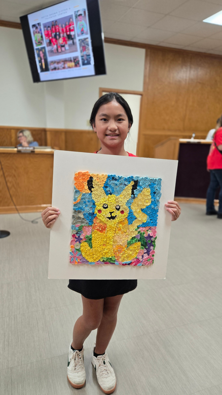 1. Student standing in the school board meeting room while holding a colorful paper-quilling artwork featuring Pikachu surrounded by flowers and a blue background.