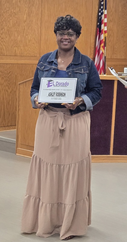 A smiling woman stands in the El Dorado School District board room holding an October 2026 Classified Employee of the Month certificate, with an American flag visible in the background.