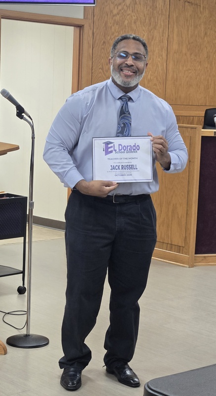 A smiling man stands in the El Dorado School District board room holding an October 2026 Teacher of the Month certificate.