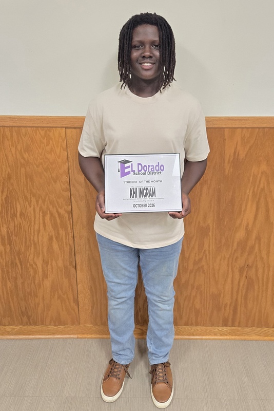 A smiling student stands in front of a wood-paneled wall holding an October 2026 El Dorado School District Student of the Month certificate.