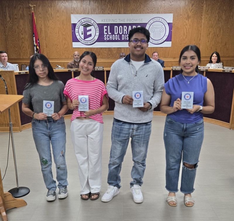 Four El Dorado High School students stand in the school board room holding Arkansas Seal of Biliteracy certificates. An El Dorado School District banner is displayed behind them as school board members sit in the background.