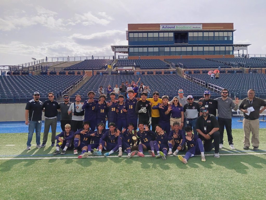 Burges High School varsity boys soccer team takes a group photo with their Regional Final trophy, Principal, Coach, Superintendent Dr. Brian Lusk, and other El Paso ISD staff and game viewers can be seen in the background