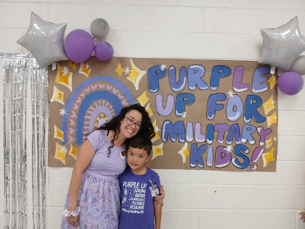 Mother and son smile for a photo while wearing purple with a purple sign behind them that says Purple Up for Military Kids