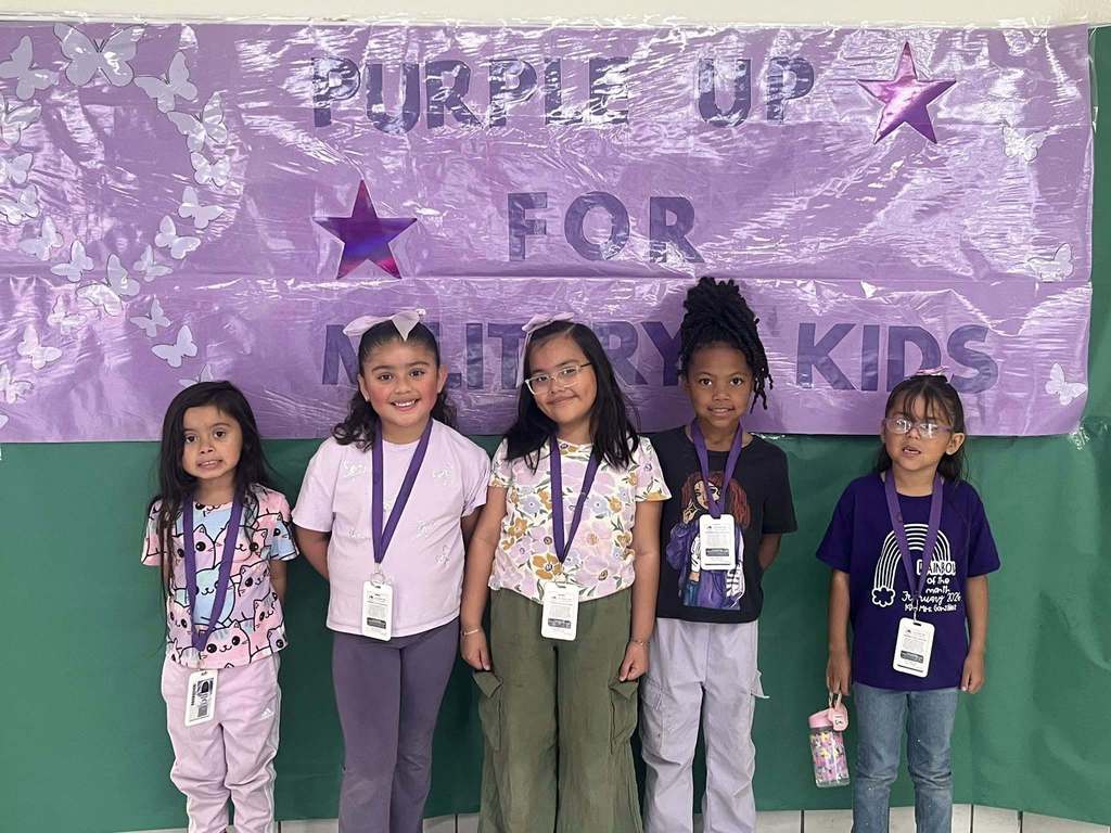 Photo of five students smiling for a group photo all wearing purple with a purple sign behind them that says Purple Up for Military Kids