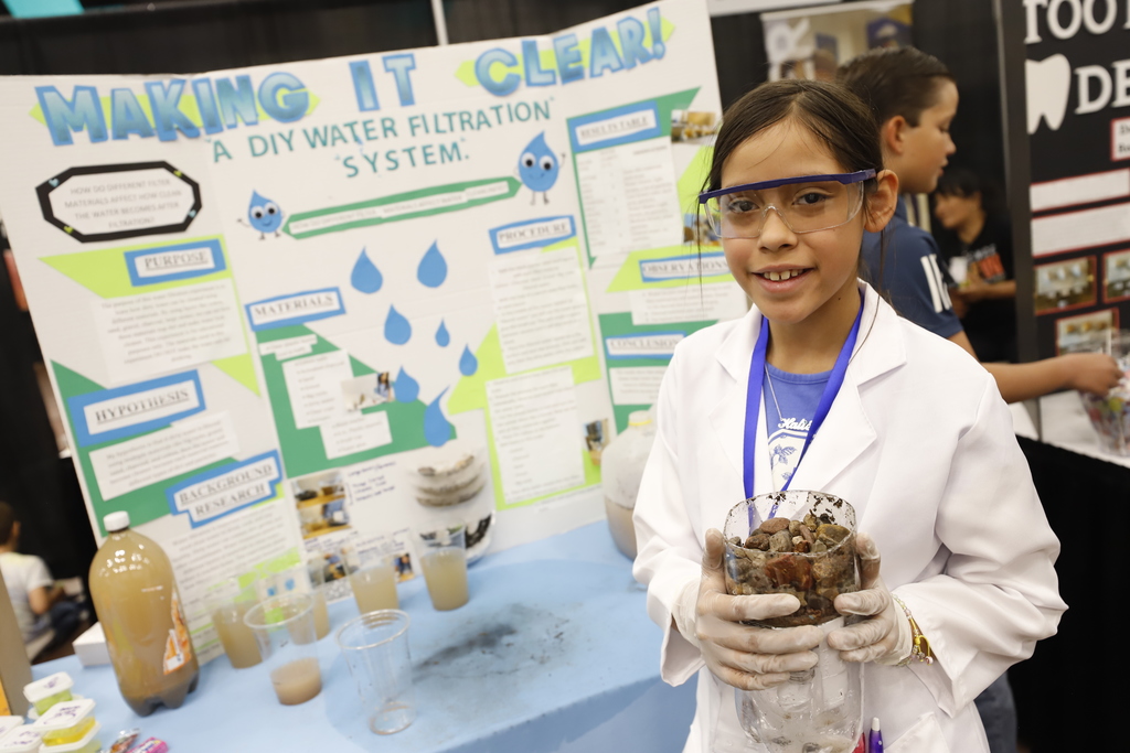Student wears goggles and lab coat while holding empty soda bottle with rocks while smiling for a photo in front of their science project that says Making It Clear! A DIY Water Filtration System