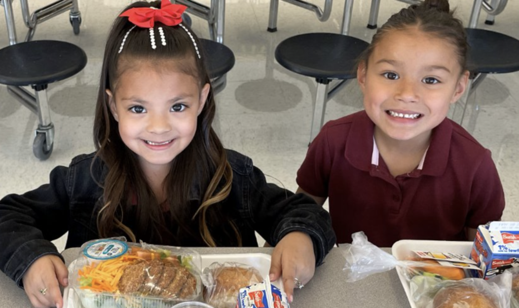 Photo of two elementary students smiling at camera with lunch meals