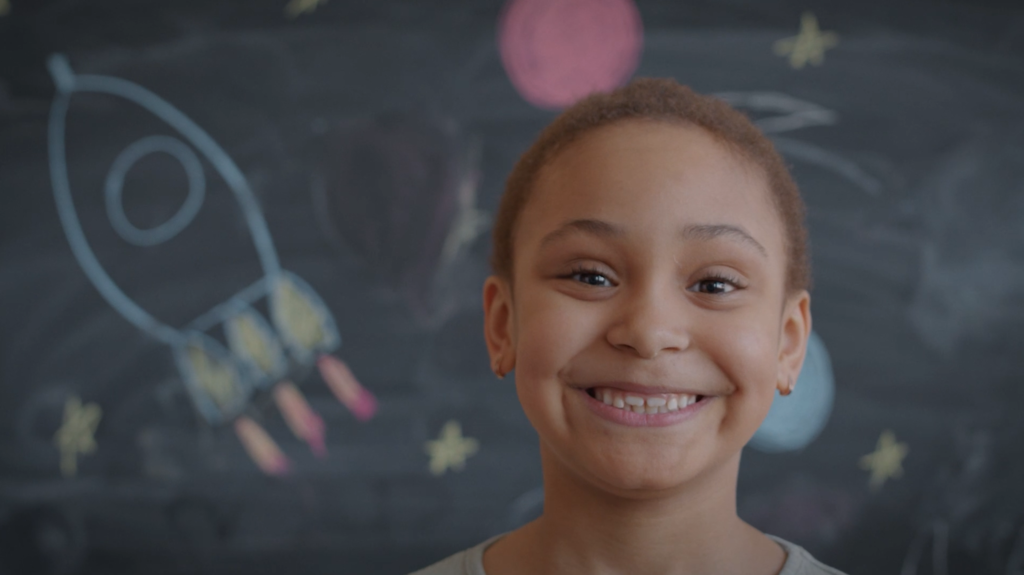 Photo of student smiling in front of chalk board with drawn rocket and stars