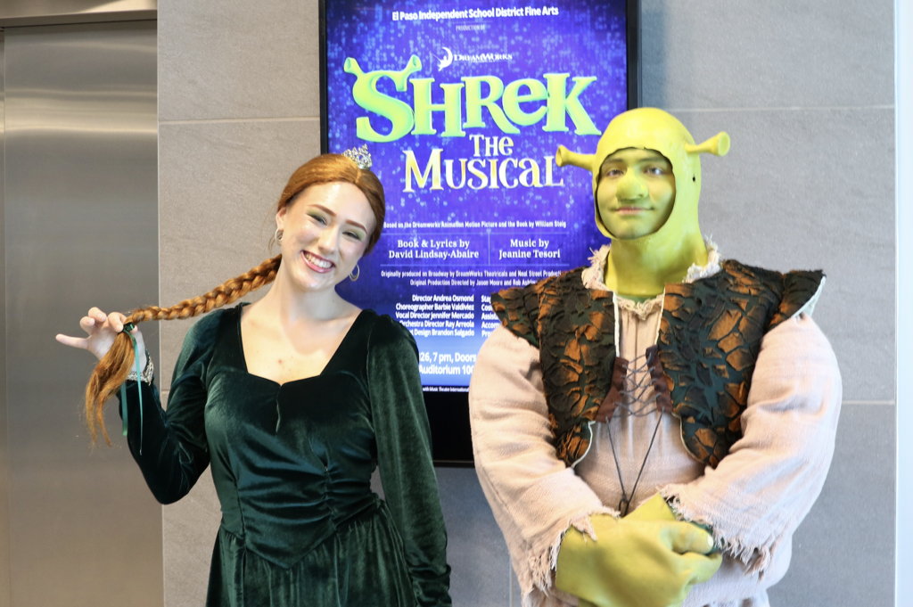 Photo of El Paso ISD Fine Arts students dressed in costume as Shrek and Fiona in front of central office screen that shows Shrek The Musical flyer