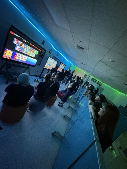 People seated in chairs facing a large screen, watching a video game. The room has blue and green lights.